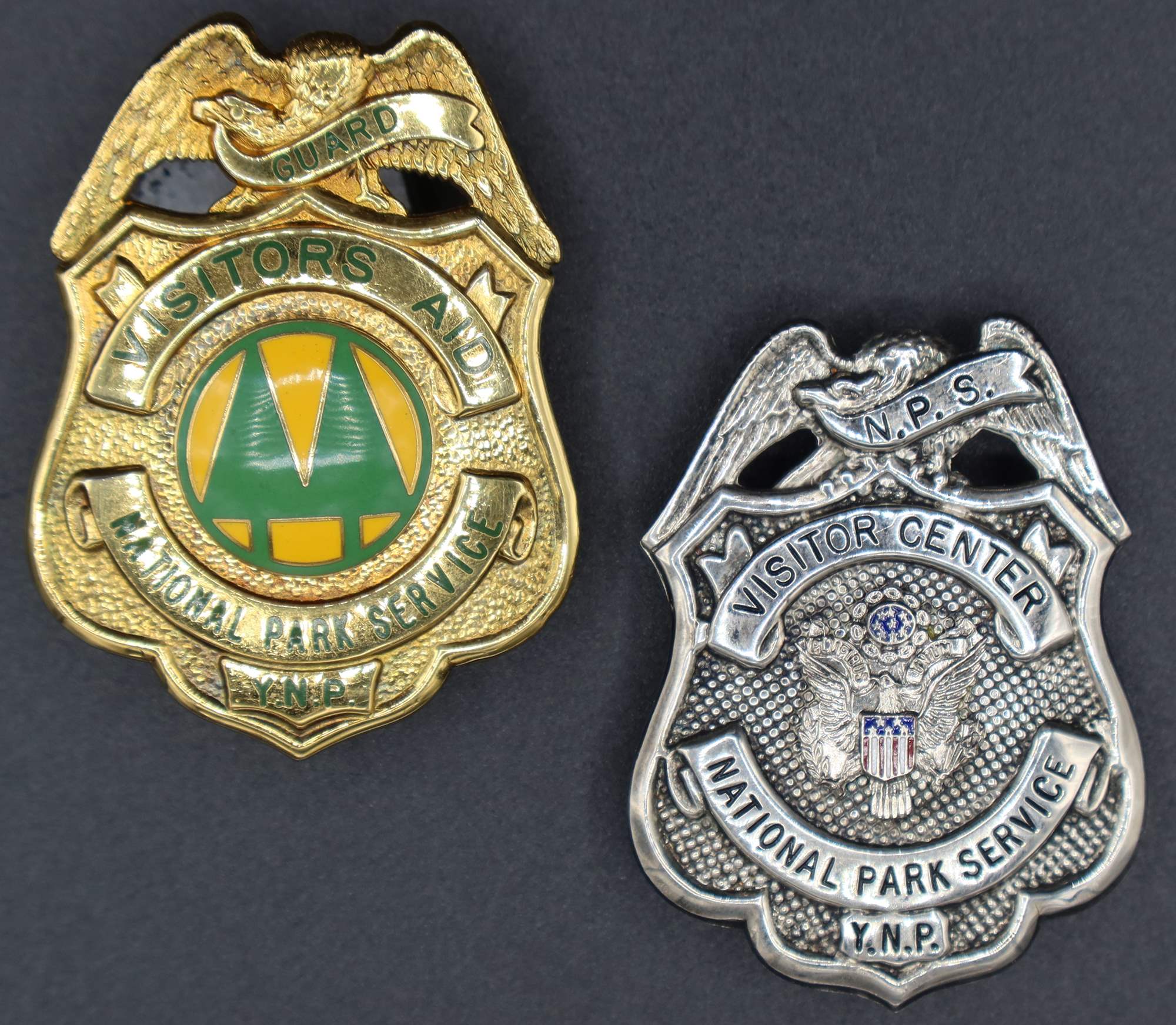 Gold visitor’s aid badge with round seal in middle with green trees on yellow. Silver Visitor Center badge with eagle with red and blue flag in middle.